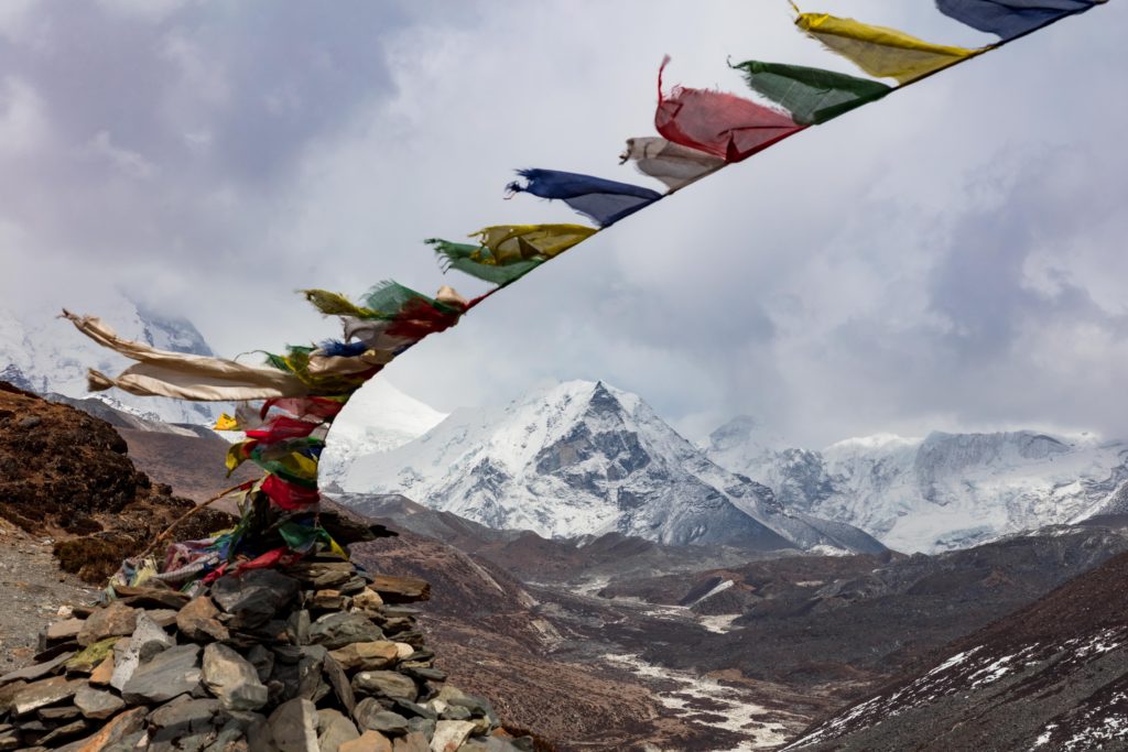 Colorful Tibetan prayer flags strung across a rocky ridge, framing snow-covered Himalayan peaks in the distance