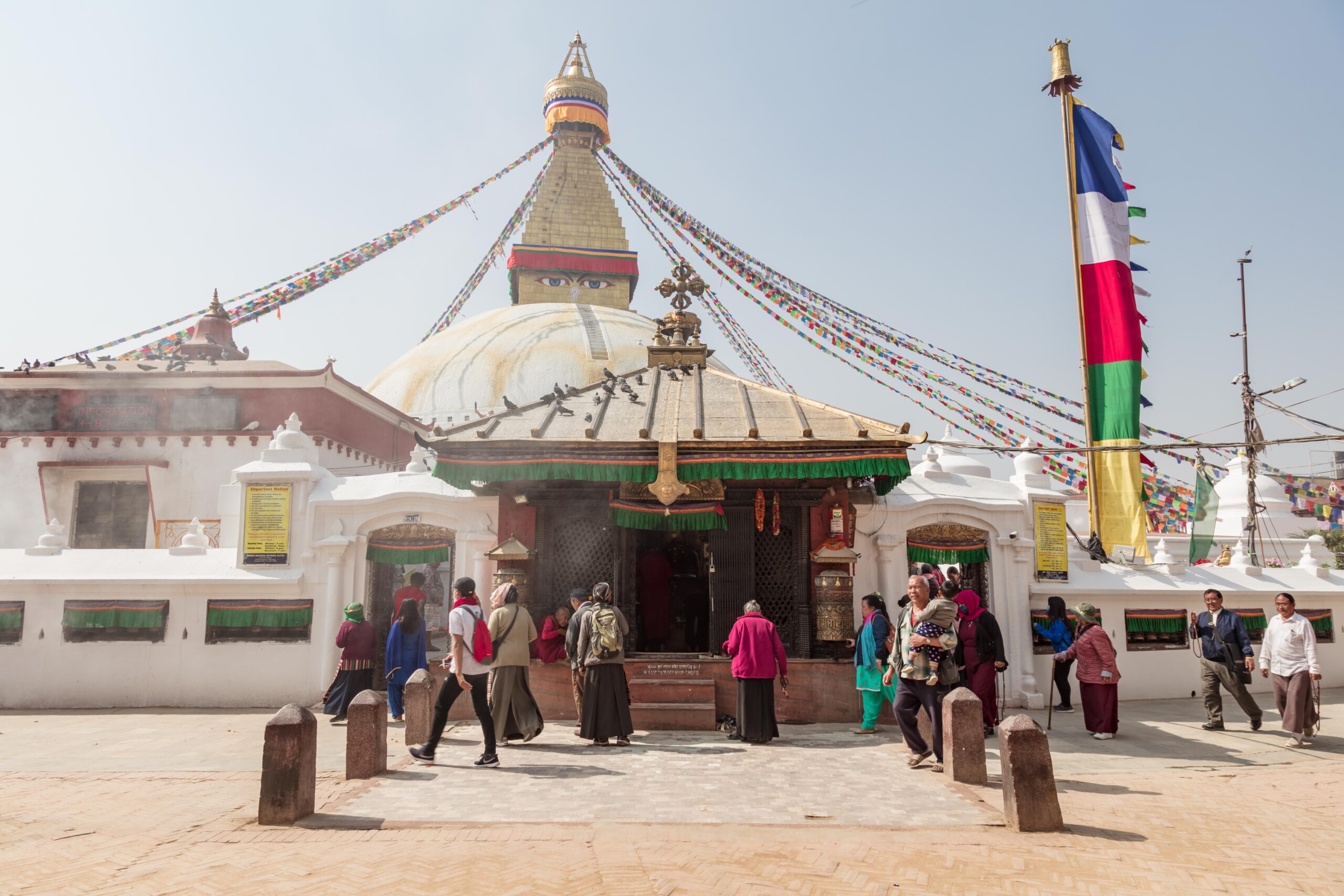 People walking and praying around Boudhanath Stupa in Kathmandu, Nepal, under clear skies with colorful prayer flags overhead