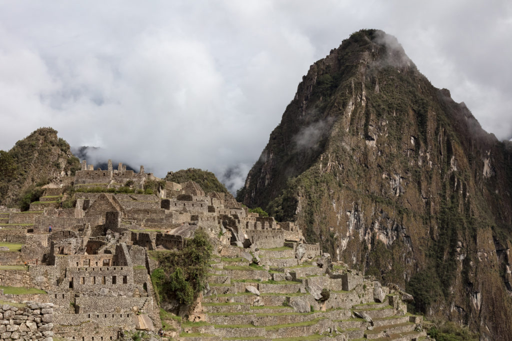 Stone ruins of Machu Picchu with terraced slopes and Huayna Picchu mountain rising behind under a cloudy sky.