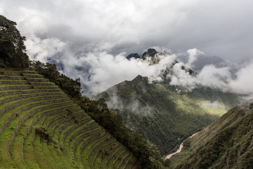 Lush green Inca terraces descend a mountainside on the left, with mist-covered Andean peaks and a winding river below.