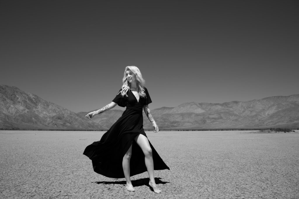 Blonde woman in a flowing black dress standing barefoot on a cracked desert floor with mountains in the background under a clear sky.