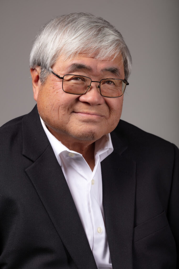 Professional headshot of an older man in a black blazer and white shirt, smiling slightly in front of a neutral gray background.
