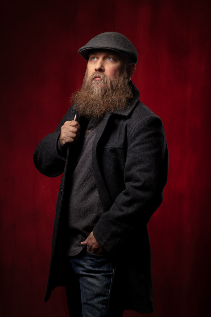Studio portrait of a bearded man in a flat cap and coat, photographed against a red backdrop with dramatic lighting — professional headshot by Brice Weaver Photography