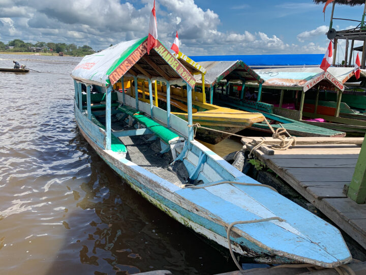 A weathered blue wooden riverboat with a canopy roof docked at the floating pier of Nanay Market in Iquitos, Peru. The boat is surrounded by other colorful local water taxis flying Peruvian flags, with the brown waters of the Amazon River in the background under a bright, partly cloudy sky.