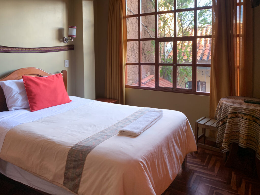 A neatly made bed with a red accent pillow in a warmly lit hotel room. A large window shows trees and traditional red-tiled rooftops of a quiet courtyard in Cusco, Peru.
