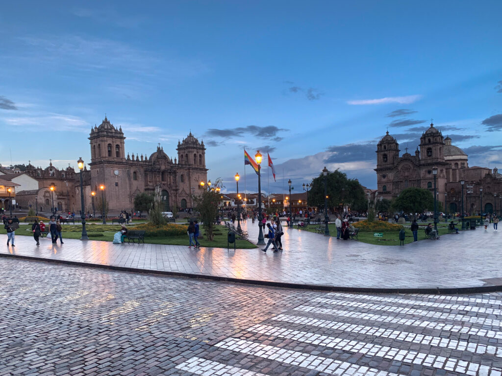 People walking across Cusco’s Plaza de Armas at dusk, with two illuminated cathedrals, cobblestone streets, and flags flying under a blue evening sky.