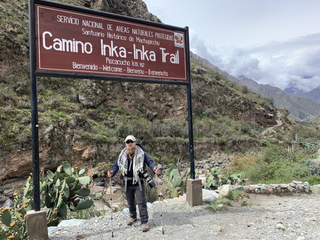 Bearded hiker with trekking poles and camera gear standing under a large “Camino Inka–Inka Trail” sign at the official starting point, surrounded by mountainous terrain and cactus plants.
