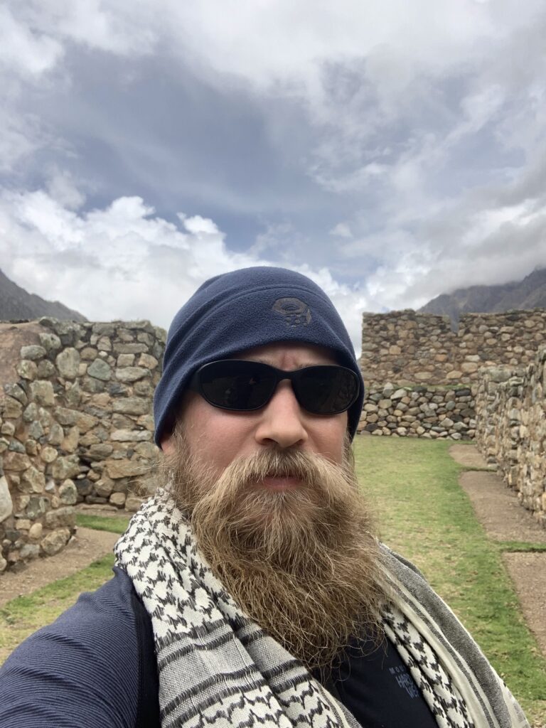 Man with a thick beard, sunglasses, and head covering takes a selfie in front of ancient Inca stone ruins with cloudy skies in the background on the Inca Trail in Peru.