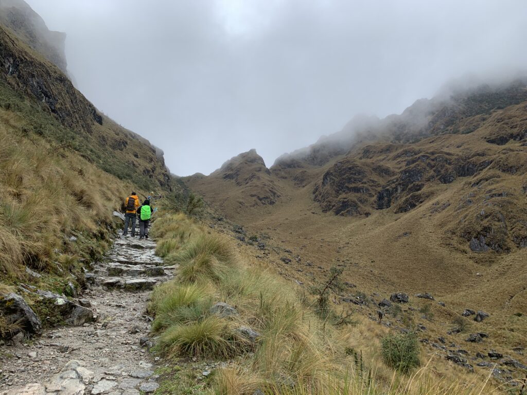 hiker ascends a misty stone path surrounded by steep grassy slopes on the Inca Trail, approaching Dead Woman’s Pass.