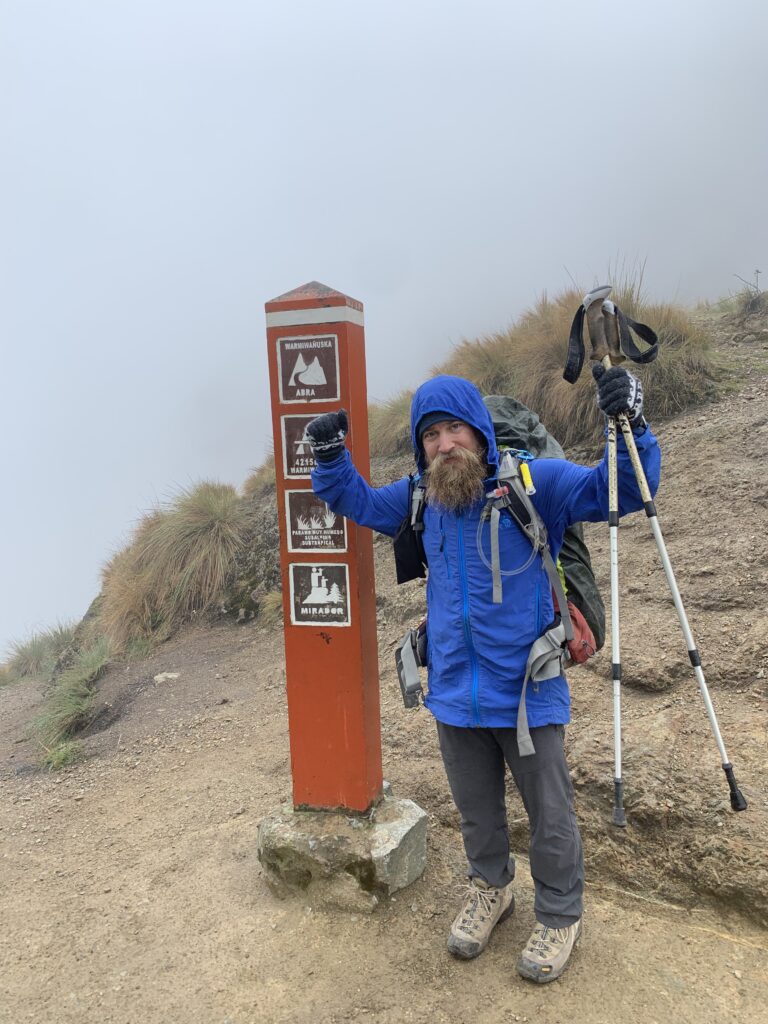 A hiker in a blue rain jacket and hiking gear poses triumphantly at the summit sign of Dead Woman’s Pass (Warmiwañusqa), the highest point on the Inca Trail at 4,215 meters, surrounded by mist and rugged terrain.