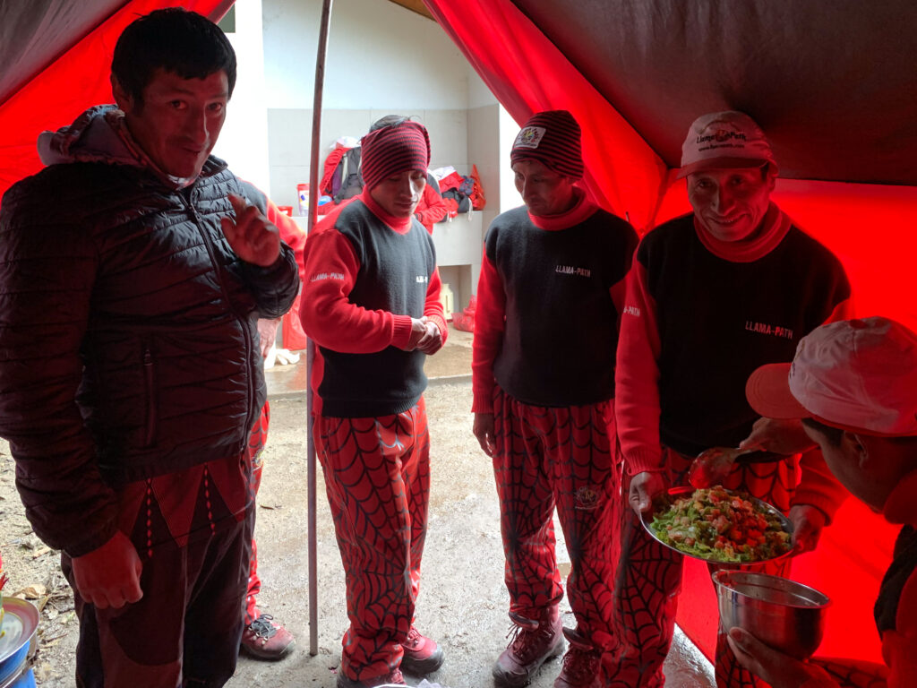 A group of Llama Path porters and chef stand inside a red cooking tent, smiling and serving a fresh salad on the Inca Trail in Peru.