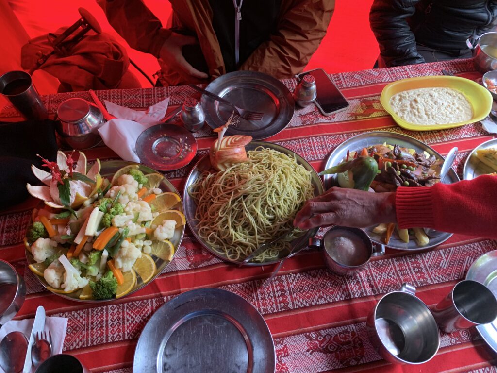 A colorful meal spread inside a red-lit dining tent on the Inca Trail, featuring plates of pasta, vegetables, stir-fry, and local dishes served on a patterned red tablecloth.
