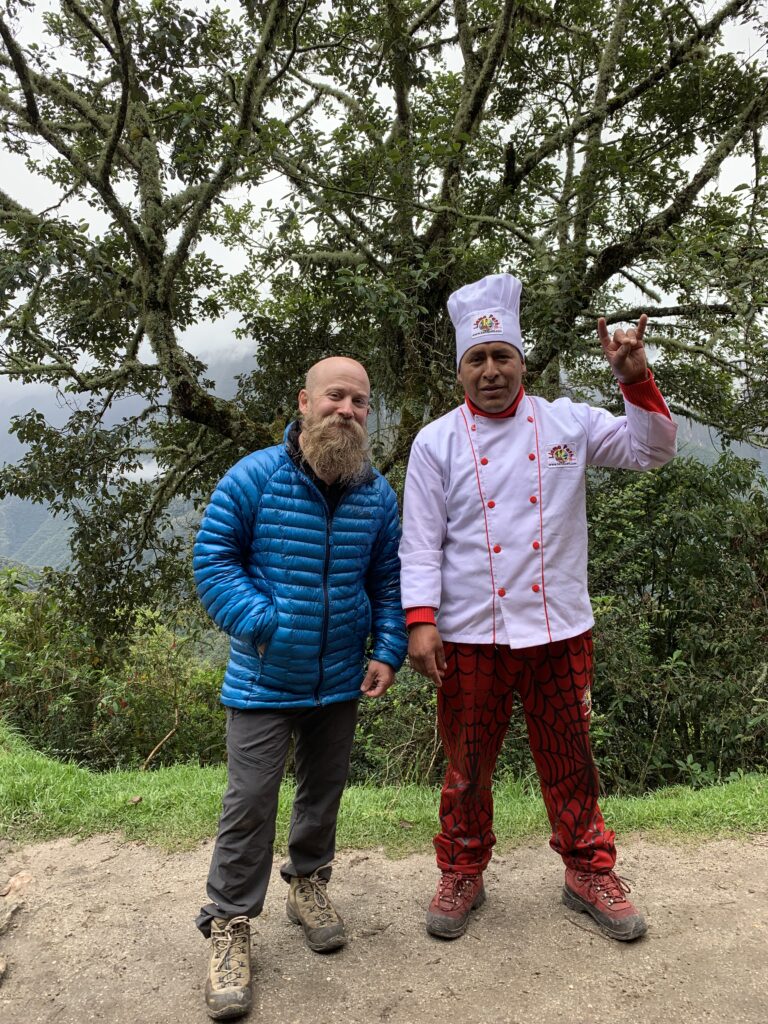 A smiling trekker in a blue puffy jacket stands next to the expedition chef in full white uniform and red spiderweb pants, posing under a mossy tree on the Inca Trail in Peru.