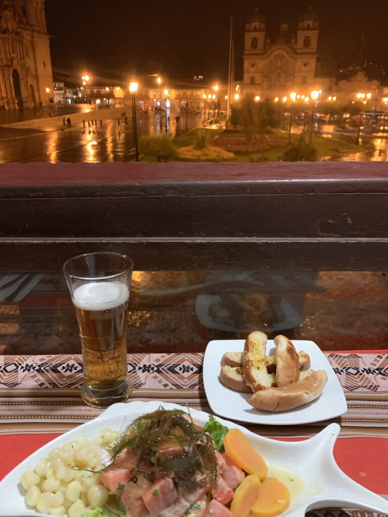 A close-up view of a table set with ceviche, toasted bread, and a glass of beer overlooking Cusco’s Plaza de Armas at night, with wet cobblestone streets reflecting warm streetlights and colonial churches in the distance.