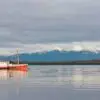 Color analog photograph of a small fishing boat on calm water along Puerto Natales Harbor in Patagonia, Chile, with snow-covered mountains in the background.